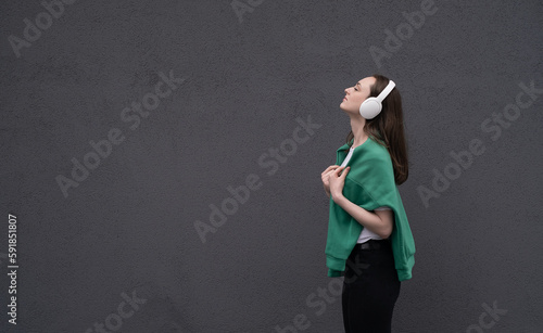 Carefree attractive brunette girl, dressed in a gray sweater, listening to music on headphones, standing on a gray background