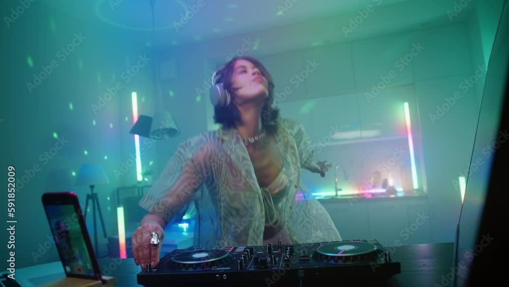 Beautiful young stylish woman dancing enjoying her live dj set in home interiors at night, excited female in headphones recording electronic music on turntable console. Colored neon lights glow