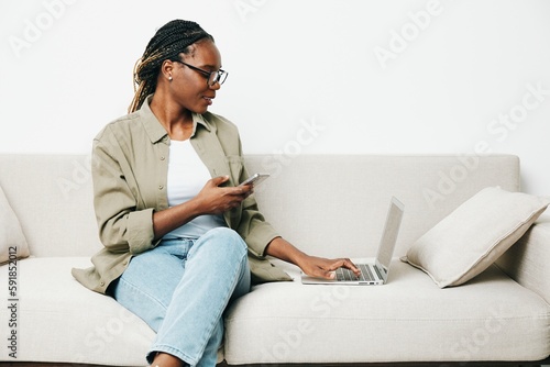 African American woman business freelancer working sitting on the couch at home in a laptop and phone, business calls and correspondence sadness and anger, home clothes and eyeglasses, light interior