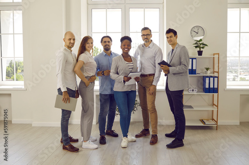 Group portrait of happy successful business people at work in the office. Team of six diverse company employees standing in a modern office and smiling. Full body length indoor shot