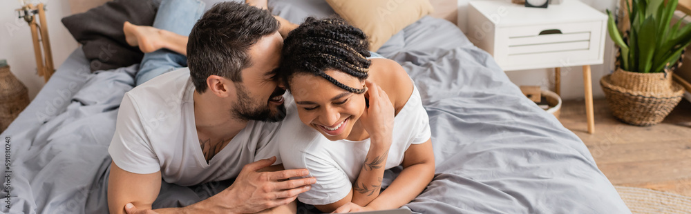 cheerful man telling secret to young african american woman while relaxing on bed at home, banner. cheerful man telling secret to young african american woman while relaxing on bed at home, banner.