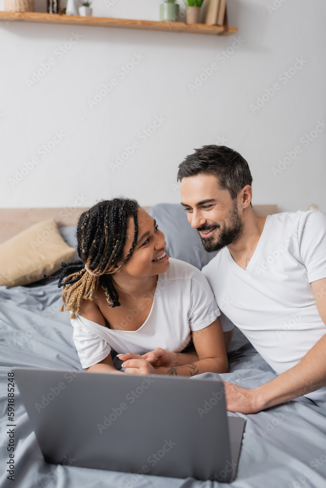 happy multiethnic couple in white t-shirts smiling at each other near laptop on bed at home. happy multiethnic couple in white t-shirts smiling at each other near laptop on bed at home.