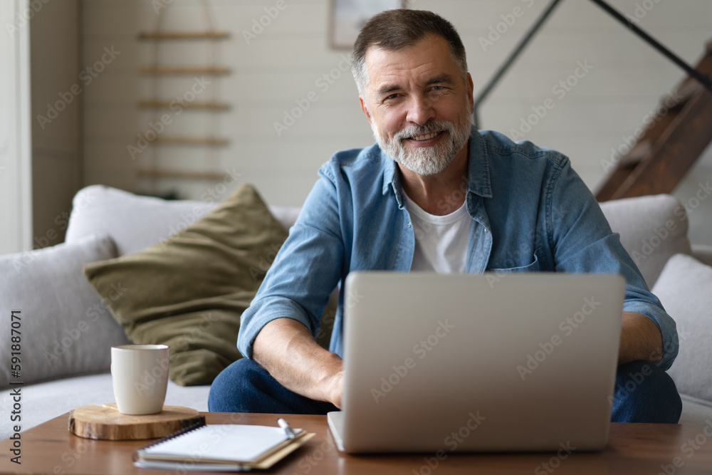 Senior man in casual clothing using laptop and smiling while sitting on the sofa, working from home. Senior man in casual clothing using laptop and smiling while sitting on the sofa, working from home.