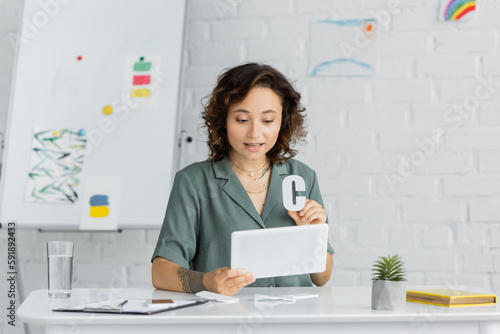 Tattooed speech therapist holding letter c during lesson on digital tablet in consulting room.