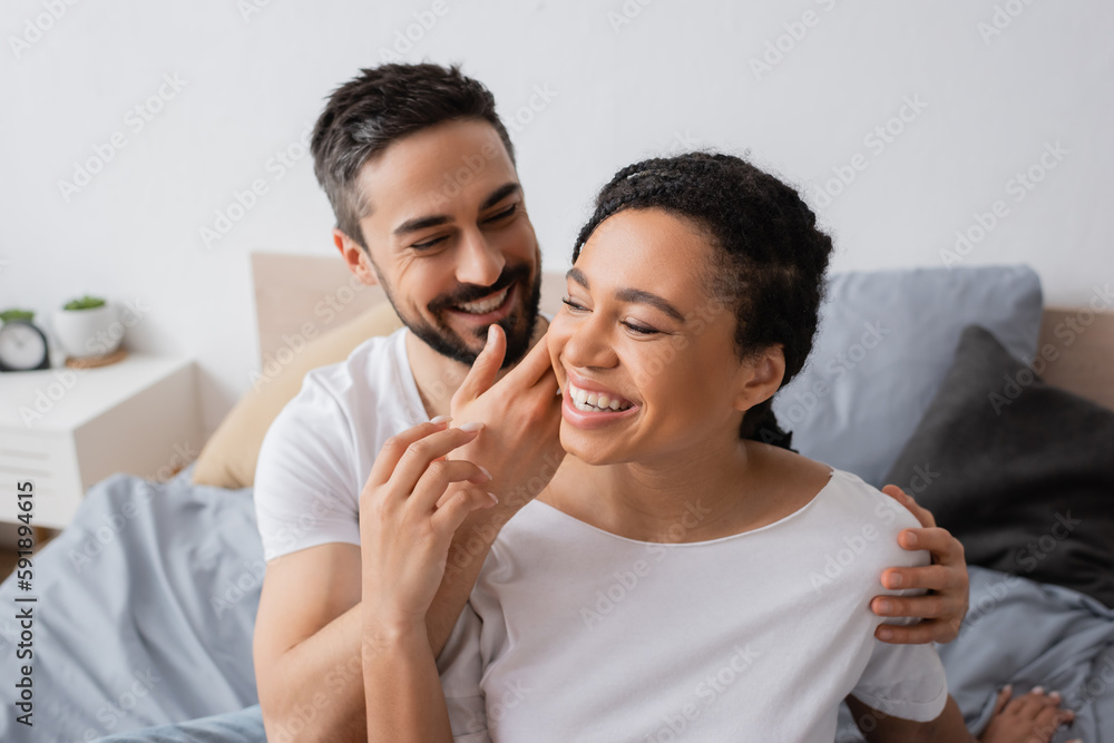smiling bearded man touching face of overjoyed african american woman in bedroom at home. smiling bearded man touching face of overjoyed african american woman in bedroom at home.