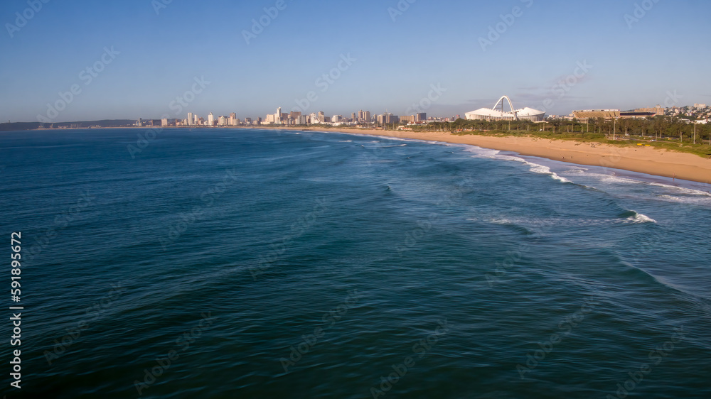 A wide view of the blue Indian Ocean and beach in Durban, with the city skyline and Moses Mabhida Stadium in the background.