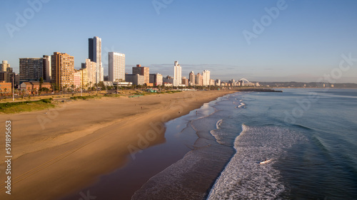 A wide view of the sea and people on Durban’s beachfront shows hotels and the city skyline with Moses Mabhida Stadium in the distance.  © Tekweni