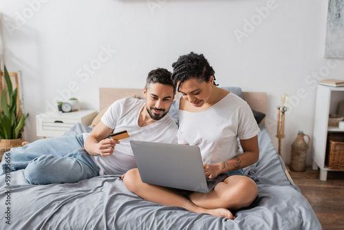 bearded man holding credit card near african american woman sitting with crossed legs and using laptop on bed at home.