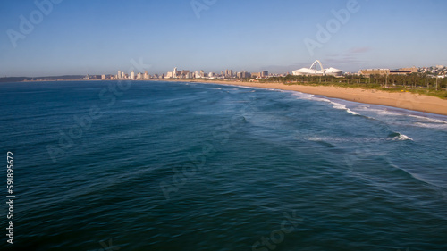 A wide view of the blue Indian Ocean and beach in Durban, with the city skyline and Moses Mabhida Stadium in the background.
