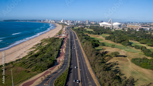A drone view of the freeway into the city of Durban, with sea and beach on one side and the golf course and the Moses Mabhida Stadium on the other. 