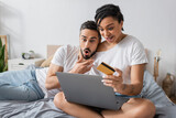 amazed african american woman with laptop and credit card near man with open mouth on bed at home.