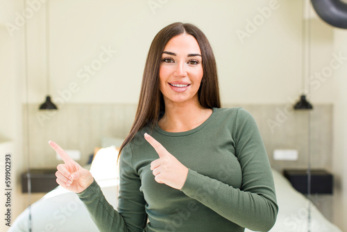 young adult pretty woman smiling happily and pointing to side and upwards with both hands showing object in copy space