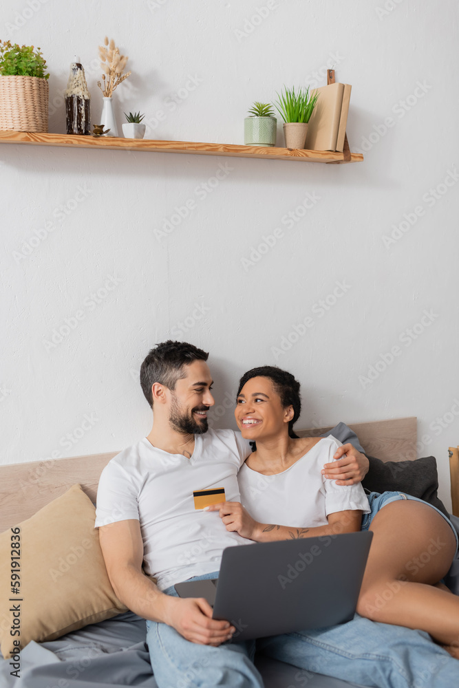 happy interracial couple with laptop and credit card looking at each other in bedroom at home. happy interracial couple with laptop and credit card looking at each other in bedroom at home.