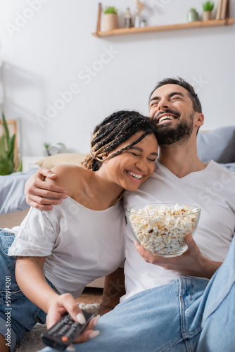 laughing bearded man with popcorn hugging smiling african american woman with tv remote controller in bedroom at home.