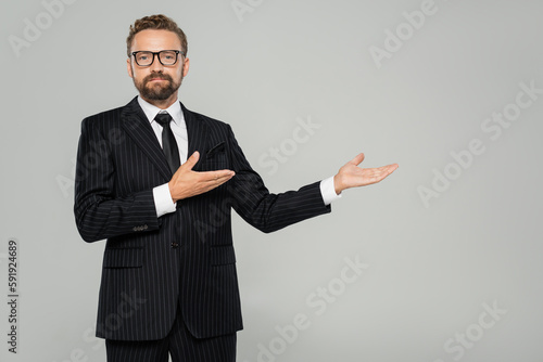 bearded businessman in formal wear and glasses pointing with hands isolated on grey.
