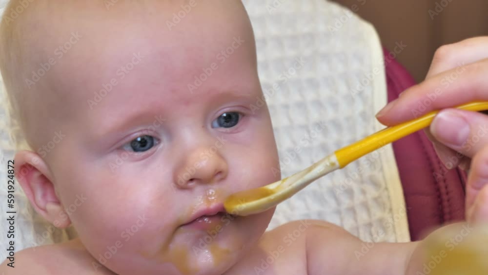 Mom feeding little boy with broccoli puree. Child at the age of six months eats broccoli while sitting on a baby chair. Slow motion shot. Mom feeding little boy with broccoli puree. Child at the age of six months eats broccoli while sitting on a baby chair. Slow motion shot.