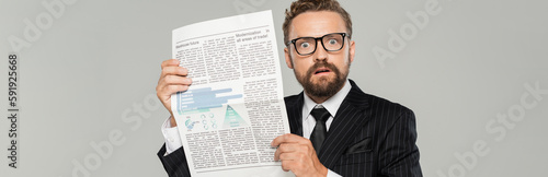 shocked businessman in suit and glasses holding newspaper isolated on grey, banner.