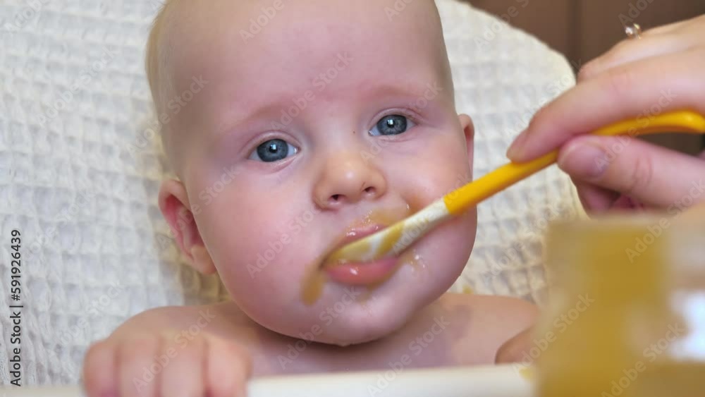 Mom feeding little boy with broccoli puree. Child at the age of six months eats broccoli while sitting on a baby chair. Slow motion shot. Mom feeding little boy with broccoli puree. Child at the age of six months eats broccoli while sitting on a baby chair. Slow motion shot.