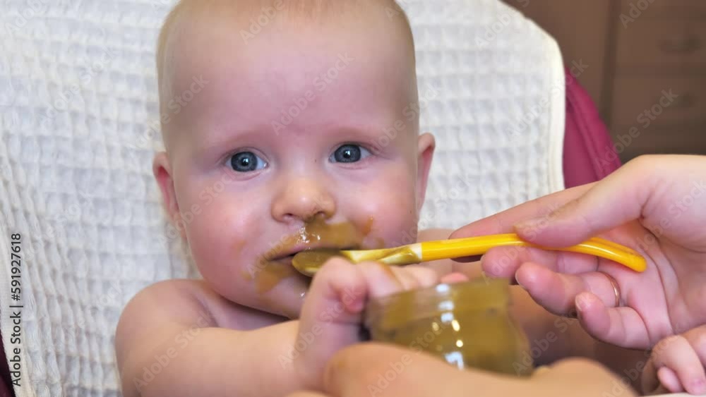Mom feeding little boy with broccoli puree. Child at the age of six months eats broccoli while sitting on a baby chair. Slow motion shot. Mom feeding little boy with broccoli puree. Child at the age of six months eats broccoli while sitting on a baby chair. Slow motion shot.