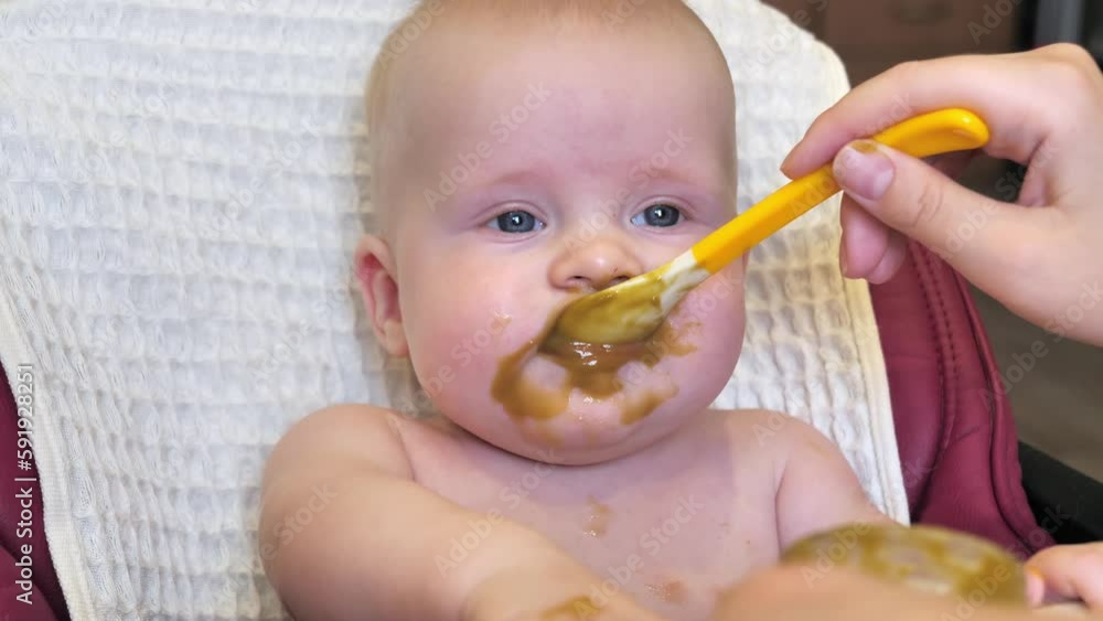 Mom feeding little boy with broccoli puree. Child at the age of six months eats broccoli while sitting on a baby chair. Slow motion shot. Mom feeding little boy with broccoli puree. Child at the age of six months eats broccoli while sitting on a baby chair. Slow motion shot.