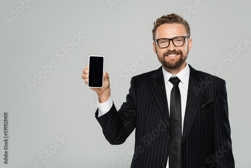 happy businessman in suit and glasses showing smartphone with blank screen isolated on grey.