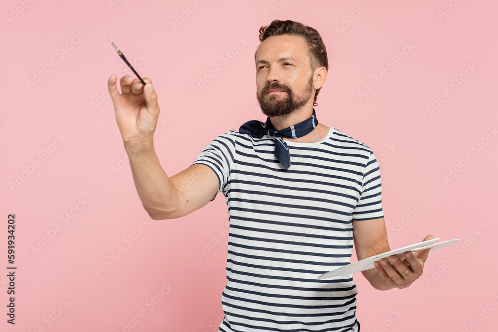 bearded french artist with neck scarf holding palette while painting isolated on pink.