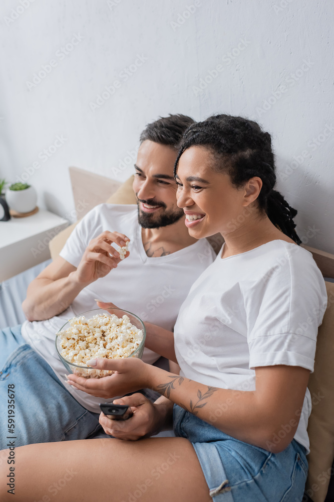 carefree multiethnic couple in white t-shirts eating popcorn and watching tv on bed at home. carefree multiethnic couple in white t-shirts eating popcorn and watching tv on bed at home.