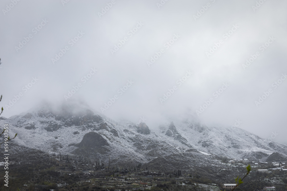 Steep rocky mountain range partially hidden by dense fog, with peaks covered in snow Steep rocky mountain range partially hidden by dense fog, with peaks covered in snow