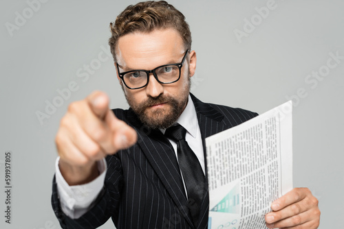 businessman in suit and glasses pointing with finger at camera while holding newspaper isolated on grey.