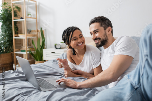 cheerful african american woman pointing at laptop and looking at smiling bearded man on bed at home.