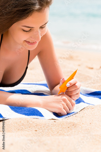 Young woman is lying on the towel at the beach and applying sunbclock from the tube on her hand