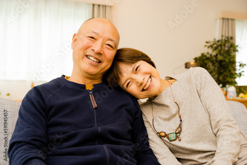 自宅のリビングで肩を預けて甘えるとても仲良しのカメラ目線の50代から60代のシニアの夫婦の笑顔 © chachamal