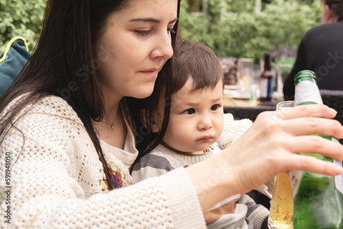 A new mum is pouring a non-alcoholic beer in her glass while holding her infant sitting in a beer garden in a pub. Spring season in Edinburgh, UK. Multicultural and multiracial family. Motherhood.