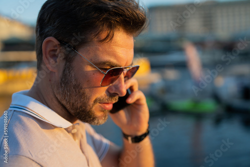 Portrait of handsome male model chatting on phone outdoor. Stylish man talking on phone dressed in polo. Fashion male posing on the street background. Business phone conversation. Urban style.