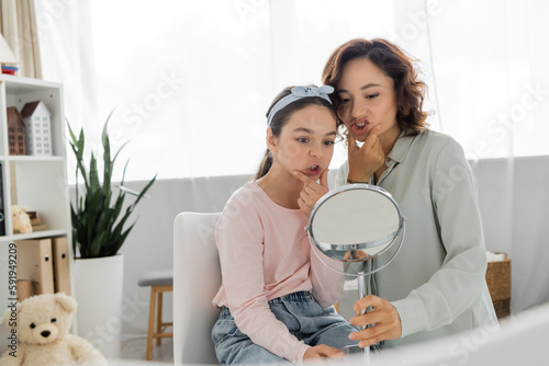 Pupil touching cheeks and looking at mirror near speech therapist in consulting room.