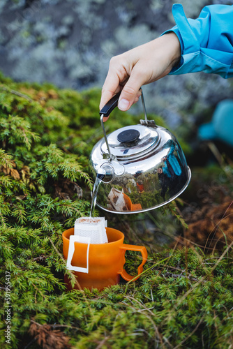 The hand holds the kettle in nature to pour boiling water into a mug, brew a coffee filter bag, an instant drink in nature, a quick preparation of breakfast.