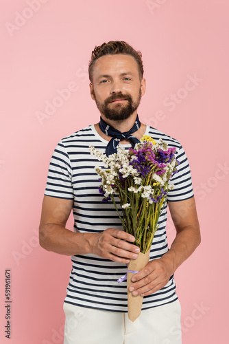 happy man in striped t-shirt and neck scarf holding flowers in paper wrap isolated on pink.