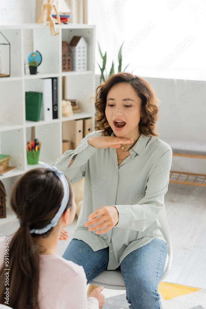 Speech therapist opening mouth and gesturing near blurred pupil in consulting room. Speech therapist opening mouth and gesturing near blurred pupil in consulting room.