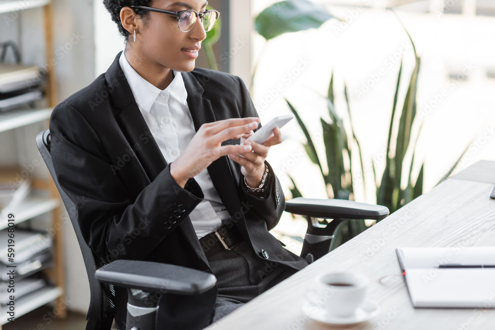 positive african american businesswoman in eyeglasses and stylish blazer messaging on mobile phone near blurred coffee cup in office. positive african american businesswoman in eyeglasses and stylish blazer messaging on mobile phone near blurred coffee cup in office.