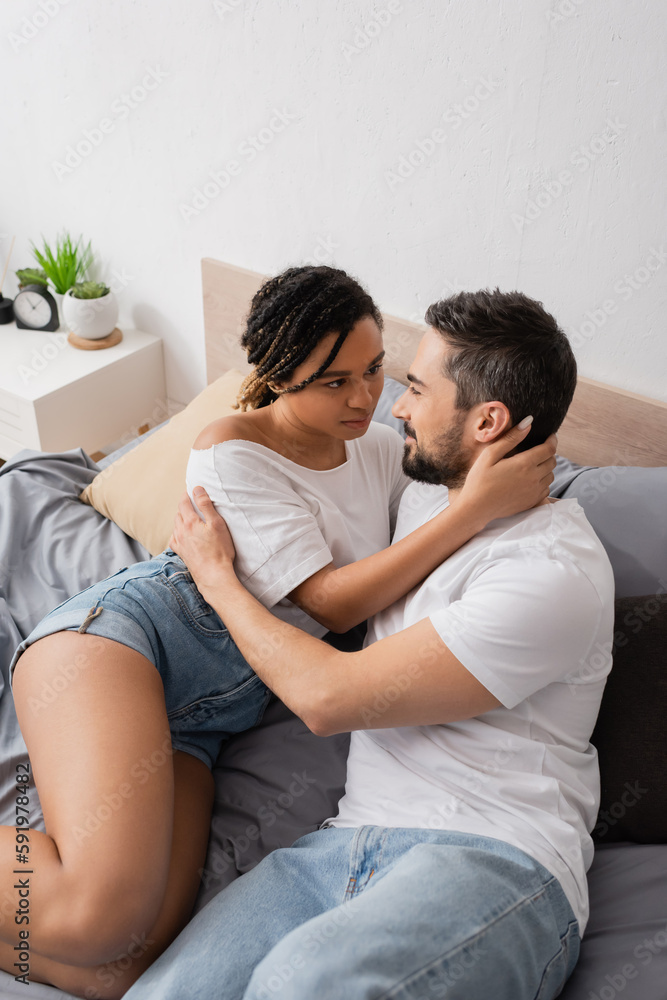 high angle view of multiracial couple in white t-shirts embracing and looking at each other on bed at home. high angle view of multiracial couple in white t-shirts embracing and looking at each other on bed at home.