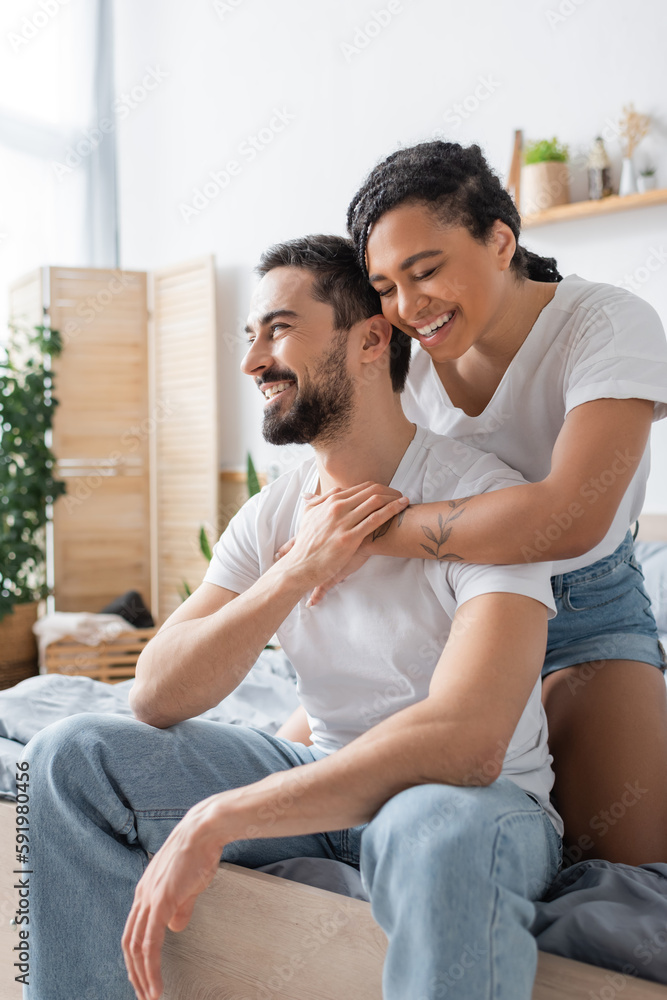 joyful african american woman with closed eyes hugging happy boyfriend in white t-shirt and jeans sitting on bed at home. joyful african american woman with closed eyes hugging happy boyfriend in white t-shirt and jeans sitting on bed at home.