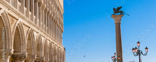 The winged lion statue atop St Mark's Column in Piazza San Marco with the white, arches of Doge's Palace (Palazzo Ducale) against a blue sky; Veneto, Venice, Italy photo