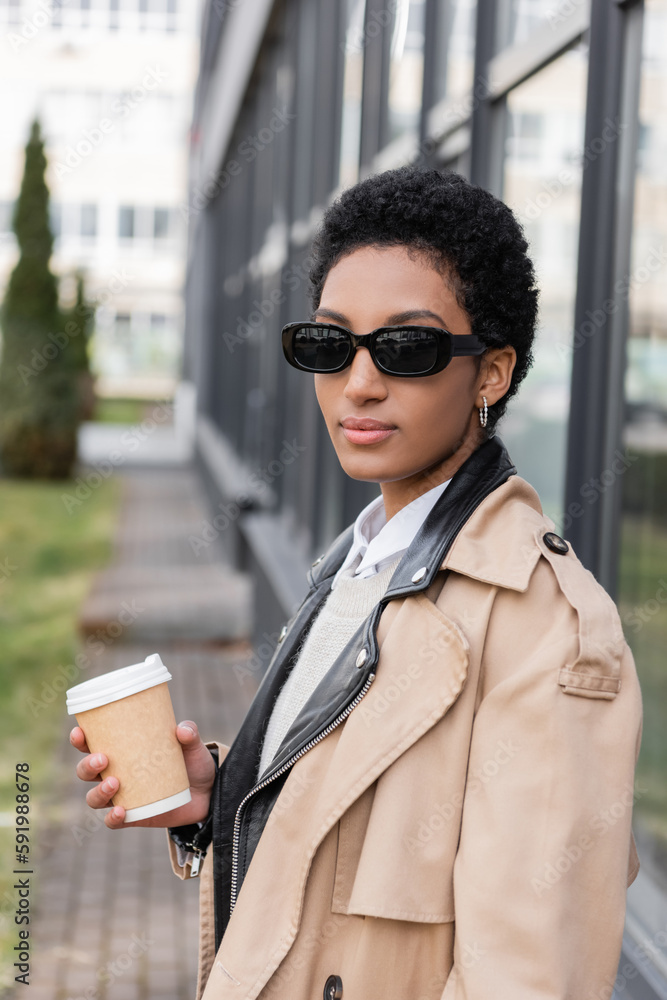 young african american businesswoman in sunglasses and trench coat holding coffee to go near blurred building on street. young african american businesswoman in sunglasses and trench coat holding coffee to go near blurred building on street.