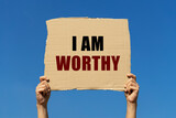 I am worthy text on box paper held by 2 hands with isolated blue sky background. This message board can be used as business concept about self worth.
