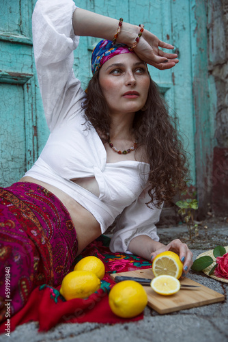 A beautiful young woman with makeup and curly hair, dressed in a colorful clothes, a gypsy,  poses outside, cuts a lemon, looking at the side. photo