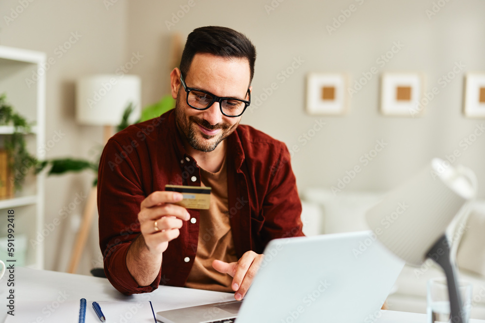 Young happy man is holding a credit card in his hand while sitting at a table with a laptop in front Young happy man is holding a credit card in his hand while sitting at a table with a laptop in front