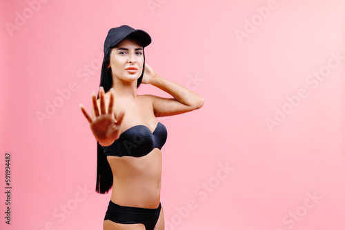 Slim woman in black swimsuit and cap showing a stop gesture with his hand and posing isolated on pink background. People summer vacation rest lifestyle concept