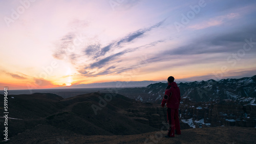 The guy in red is walking to the edge of the canyon. Red-orange light from the rising sun. Purple clouds. Orange canyon Charyn. Morning. There is snow in places. A trip through the canyon. Kazakhstan