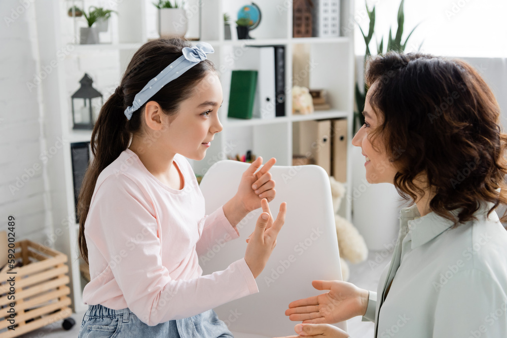 Side view of preteen girl gesturing and talking to smiling speech therapist in consulting room. Side view of preteen girl gesturing and talking to smiling speech therapist in consulting room.