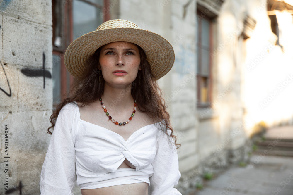 Closeup portrait of a beautiful brunette with curly hair, wearing a straw hat and a white blouse and skirt, poses outside. Copy space.	
